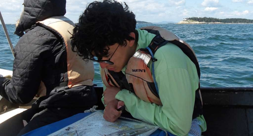 A student wearing a life jacket sits on a sailboat and examines a map. 